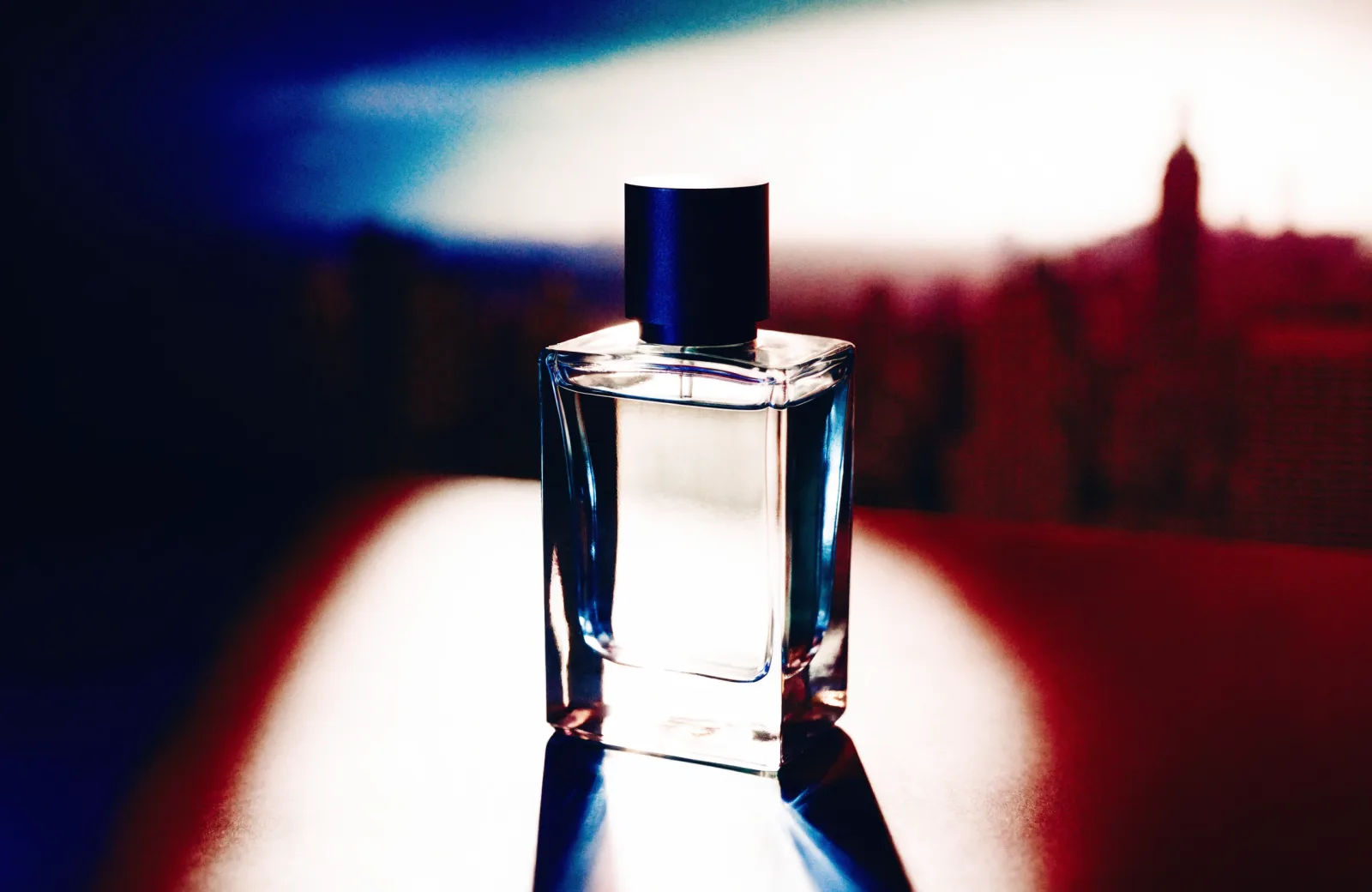 A clear square perfume flacon with a matte black cap, lit against a dark editorial backdrop